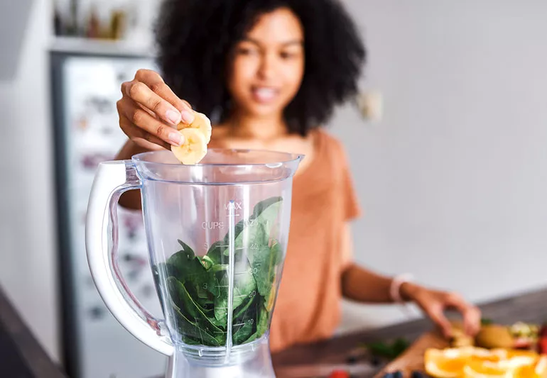 Woman adding banana to blender with spinach for healthy smoothie