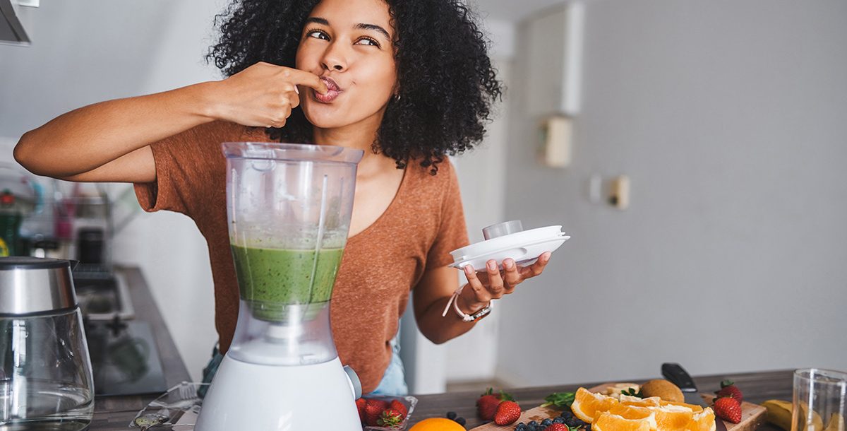Woman tasting a fresh green smoothie from a blender in a bright modern kitchen