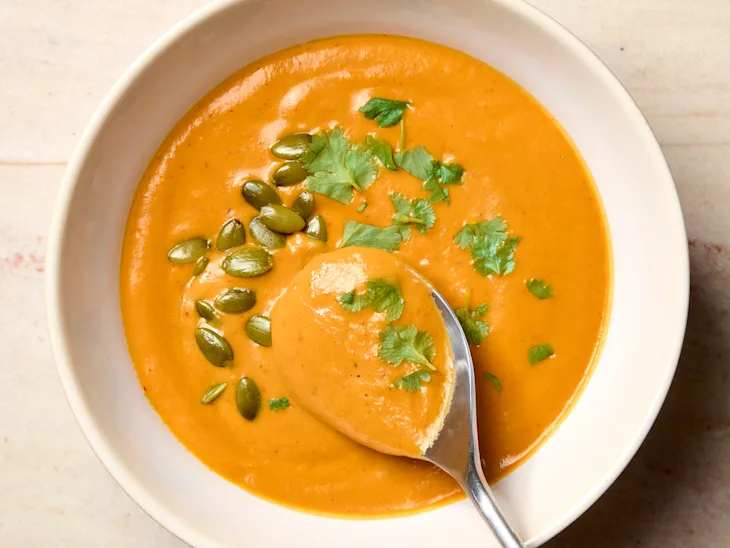 Close-up of creamy pumpkin soup in a bowl with a spoon, garnished with pumpkin seeds and fresh herbs
