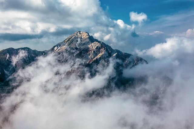 Misty Mountain Peak A dramatic mountain summit piercing through thick clouds, with rugged rocky faces partially obscured by swirling mist, creating an ethereal and majestic alpine atmosphere.