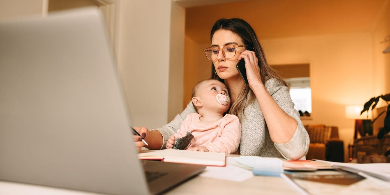 Mother working on laptop while holding baby in home office setting