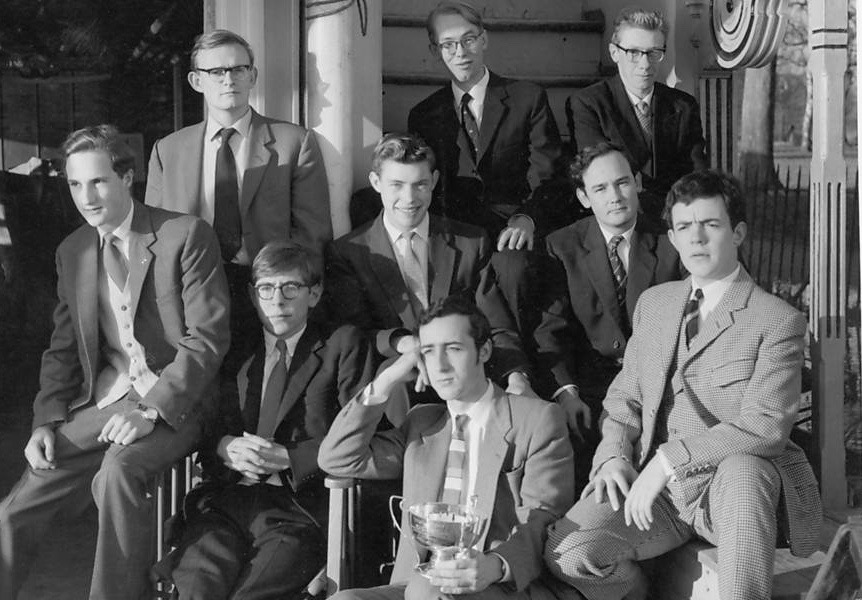 Foto em preto e branco de Stephen Hawking jovem sentado entre colegas de Oxford, todos de terno