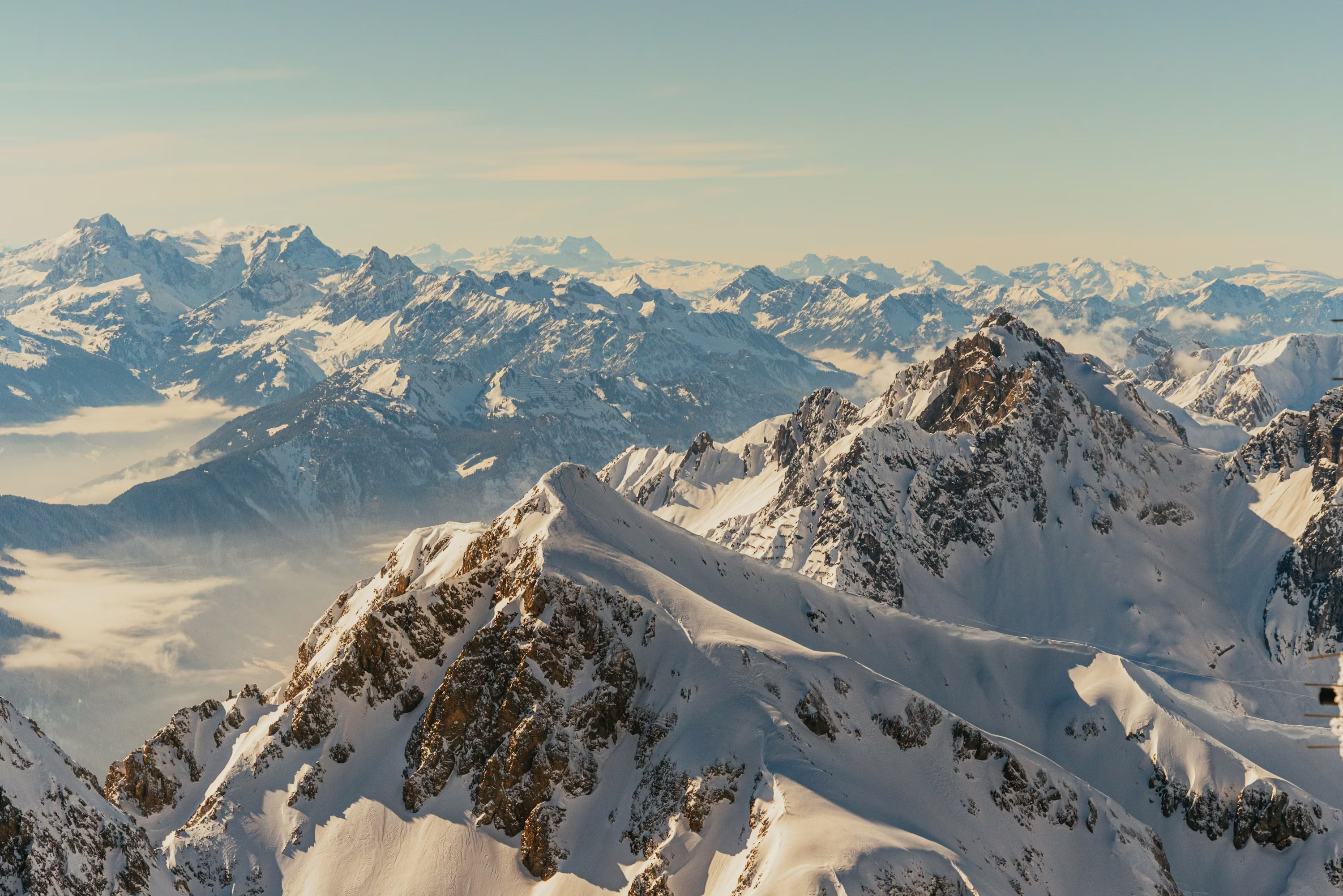 An expansive panorama of snow-covered mountain peaks stretching to the horizon, bathed in soft golden light, with layers of ridges fading into the distance above a sea of clouds.