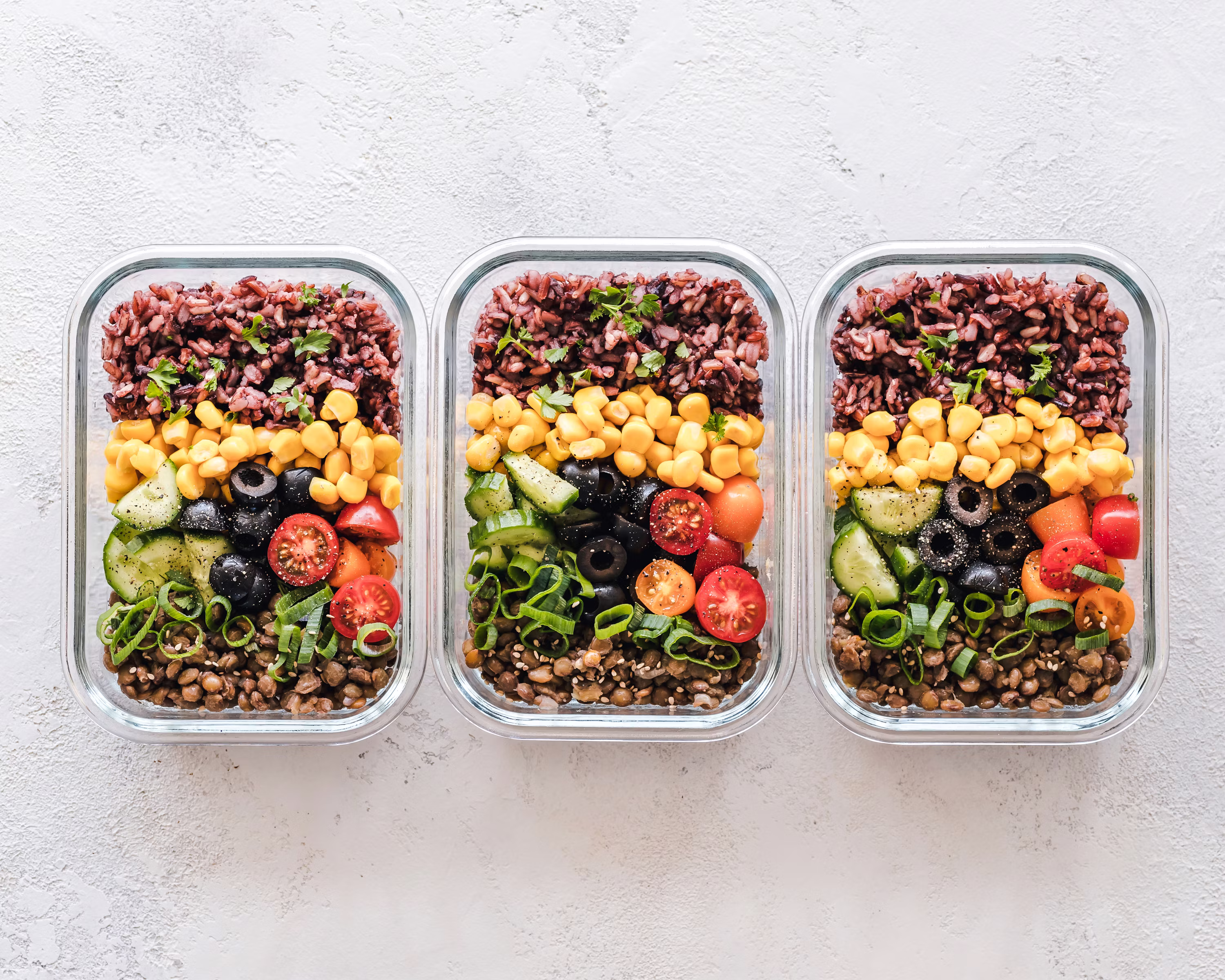 Three colorful meal prep containers with rice lentils corn tomatoes cucumbers