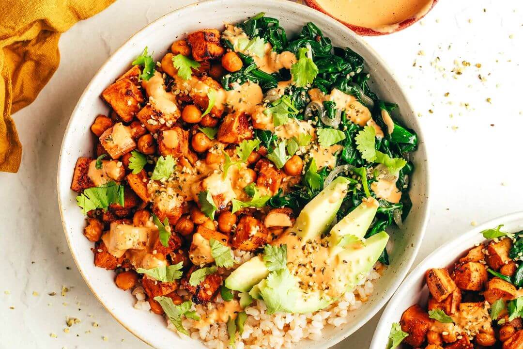 Colorful healthy grain bowl with chickpeas, roasted sweet potato, kale, avocado, and tahini dressing