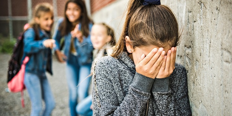 Estudante adolescente vítima de bullying escolar, com expressão de tristeza, enquanto outras estudantes conversam ao fundo