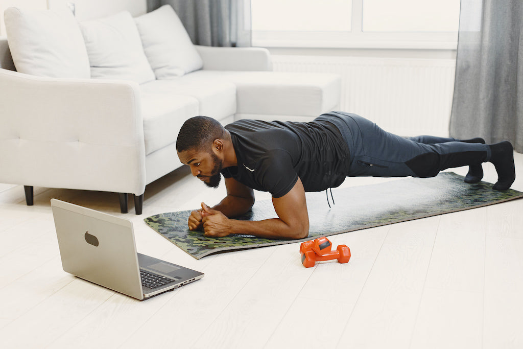 Man doing plank exercise on yoga mat at home with laptop nearby