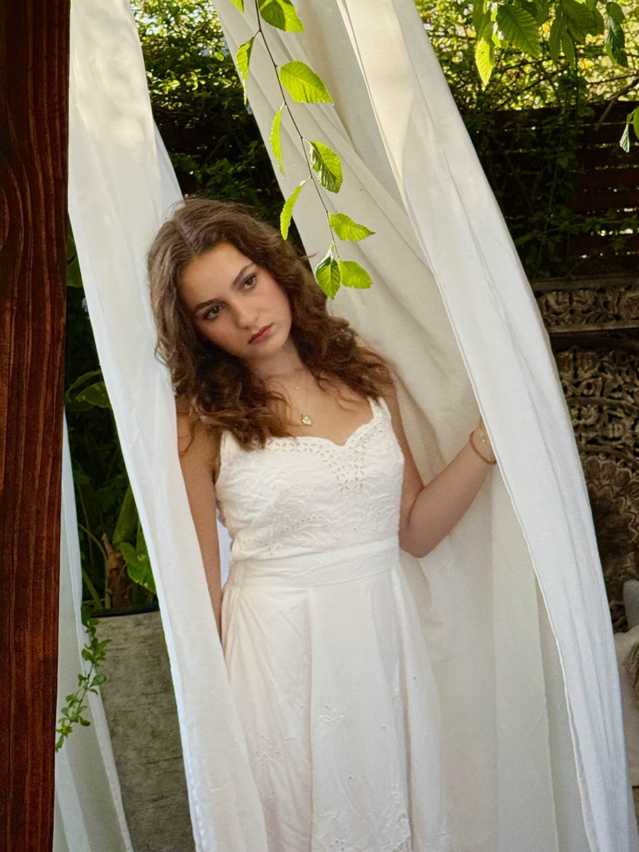 Ellory Grace in a white lace dress standing between flowing white curtains with greenery