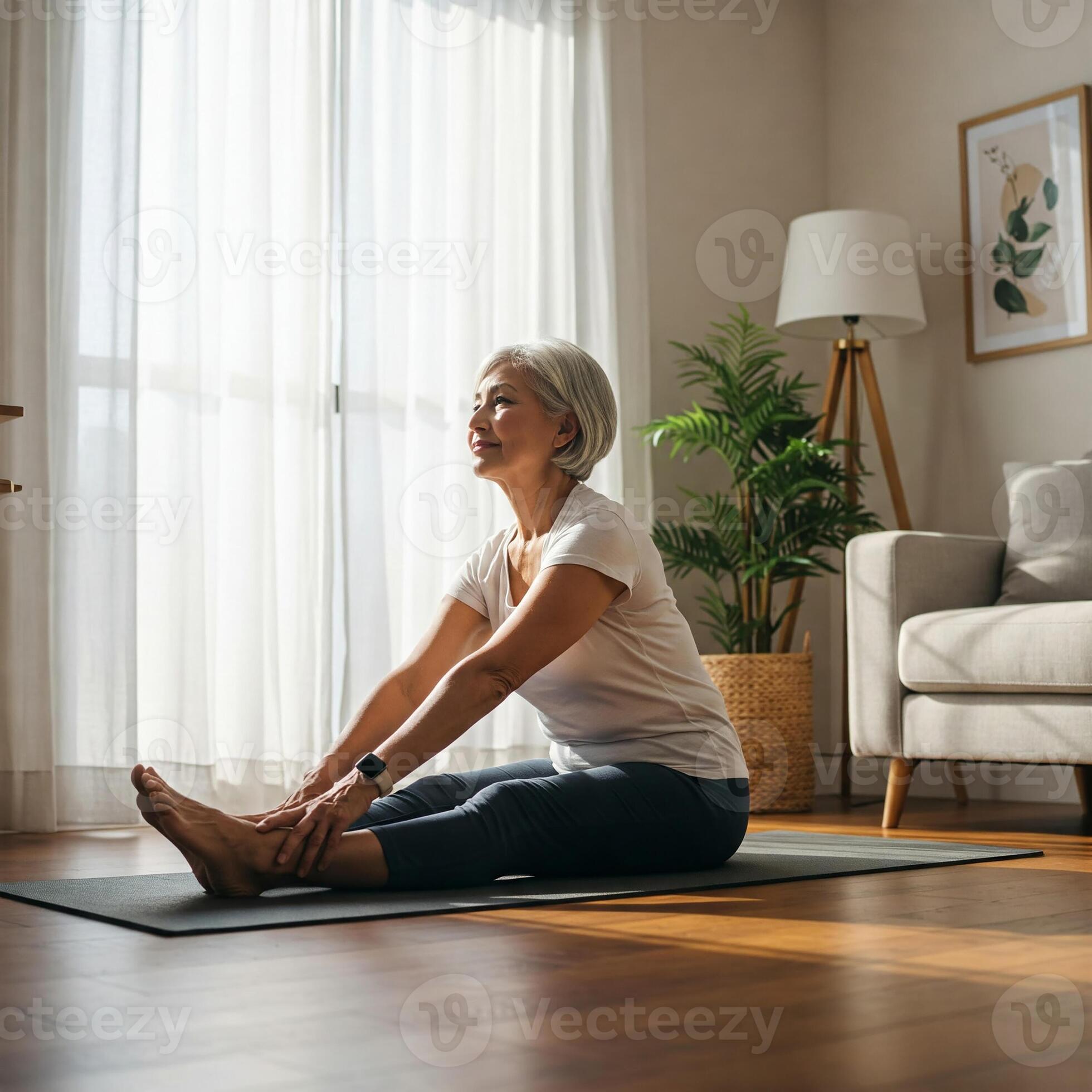 Senior woman practicing yoga at home in bright living modern room. She sits on mat doing side stretch, smiling and enjoying healthy lifestyle. Peaceful morning wellness routine with natural light 60189012 Stock
