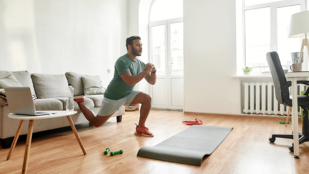 Beginner home workout in living room with no equipment needed Man doing lunges in a bright home living room with yoga mat and laptop