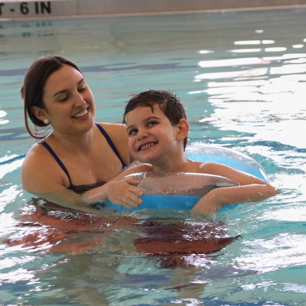 Professional swim instructor providing gentle support to a smiling child with special needs in a warm pool environment