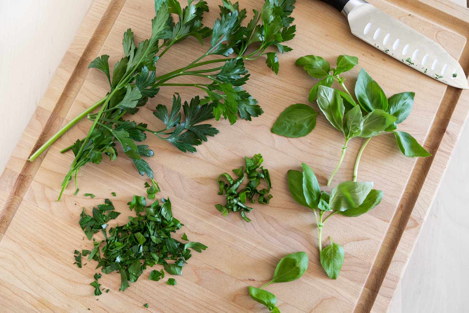 Fresh parsley and basil being chopped on a wooden cutting board with a chef's knife