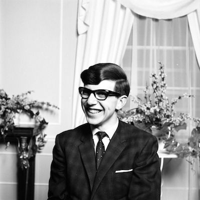 Foto em preto e branco de Stephen Hawking jovem, sorrindo, usando óculos e terno